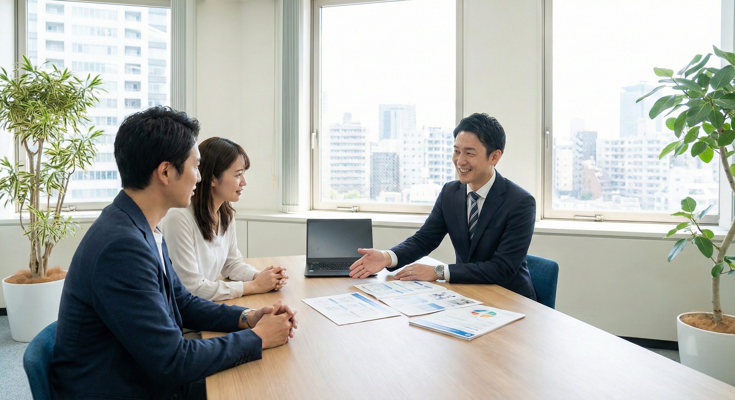 明るい会議室でクライアントと対面で商談している営業担当者の写真。テーブルの上にはノートPCと提案資料が広げられている。